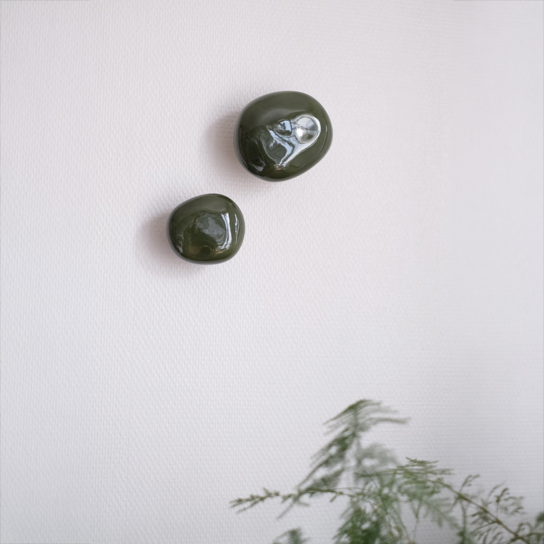 Deux patères Agate vert mousse en porcelaine émaillée, installées sur un mur blanc et vue de face #couleur_vert mousse